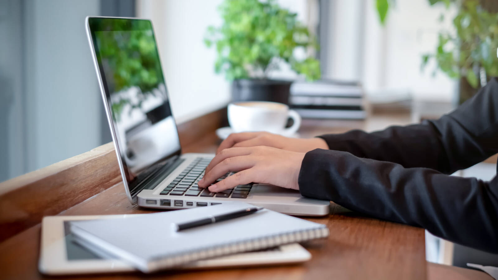 A professional seated at a wooden desk types on a laptop in a clean, well-lit workspace. The scene includes a neatly arranged notebook with a pen, a digital tablet, and a cup of coffee in the background, creating a focused and organised environment. Green potted plants add a touch of natural decor, while the soft daylight enhances the calm and productive atmosphere. The individual is dressed in business attire, suggesting remote work, independent project work, or virtual collaboration in a modern business setting.