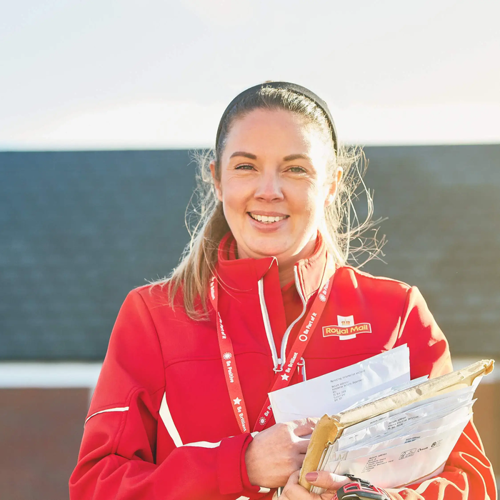 A Royal Mail postal worker wearing a red uniform smiles while holding letters and small parcels outside residential homes.
