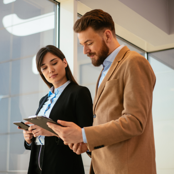 Two business professionals, a woman and a man, stand near a large window. They are both wearing business attire; the woman has on a dark-colored blazer and the man has a light beige or tan blazer. Both appear to be looking at and holding digital tablets or clipboards. The woman is looking down at the tablet or clipboard in her hands, and the man is looking at the tablet/clipboard in front of him while glancing over at the woman. The building exterior, visible through the window, reflects a light blue sky. The background suggests an interior office space.