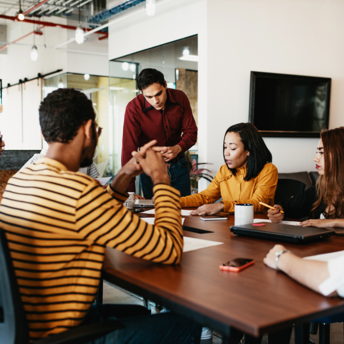 This image captures a collaborative team meeting in a modern office setting, where colleagues are gathered around a table engaged in discussion and problem-solving. One individual stands to review or explain key points while others listen attentively, take notes, and contribute ideas. The workspace features a contemporary design with glass walls, exposed ceilings, and warm lighting, creating an open and professional atmosphere that encourages teamwork and productivity.