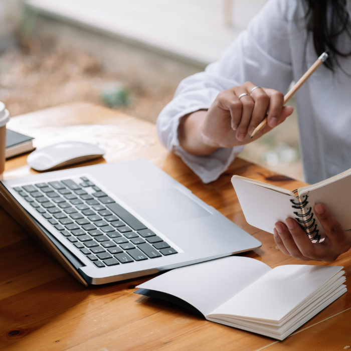 A person sits at a wooden table, holding a spiral-bound notebook and a pencil, looking intently at the open notebook.

The scene is composed of a close-up view of a person's hands holding a notebook and a pencil. The person appears to be focused on the task of writing or reading.  The notebook is positioned centrally, with hands holding it open, and a computer laptop lies on the table beside it. The lighting is soft and ambient, highlighting the person's hands and the notebook. The wooden table serves as a neutral backdrop, and the focus is squarely on the person's actions and the notebook.

The primary subject is a person, likely an adult, whose hands and upper body are visible. The person is holding a spiral-bound notebook and a pencil, appearing to be deeply engaged in reading or writing.  The person's attire is a light-colored, likely white or beige, long-sleeved shirt, and the hands and upper arm portion of the person are visible.  No other details of the person are visible beyond the hands and upper torso.

The artistic medium is photography. The technical execution is skillful, with the image focusing on a soft focus on the notebook, and hands holding it. The style is straightforward, naturalistic, and simple, capturing a casual moment of focus.  The creative choices emphasize the quiet focus of the person engaged in their work. The focus is on the moment and simple details, without artistic embellishments or special effects.

The setting is a simple, wooden table, likely indoors. The lighting conditions are soft and natural, casting no harsh shadows. The environment appears to be a workspace or home office, providing a sense of calmness and productivity. The wooden surface and soft lighting contribute to the overall atmosphere. The setting is uncluttered, allowing the focus to remain on the person and the activity.