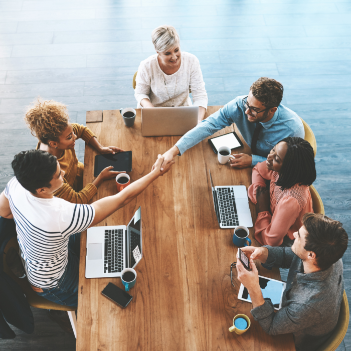 A high-angle shot captures a group of diverse people seated around a wooden table, engaged in a business meeting or collaboration session, with a handshake taking place in the center.

The scene composition is a top-down view of a rectangular table where several people are seated, creating a circular arrangement around the table.  Hands are outstretched in a handshake, signifying a collaborative or agreement-making moment. The table is filled with laptops, coffee cups, and other digital devices, suggesting a workplace setting. The positioning of the individuals around the table creates a sense of focused interaction and collaboration. The framing is tight, centering the action of the handshake, highlighting the group's interaction and activity.

The key subjects are various adults, comprising a diverse group in terms of gender and ethnicity. Their attire suggests a professional or business context. Facial expressions are varied but generally show engagement and attention. One particular interaction of the people is the handshake, and the overall mood seems collaborative and positive.

The artistic medium is photography, capturing a candid moment in a contemporary office setting.  The technical execution is clear, showing sharp focus on the subjects and the table, showcasing a professional quality image. The style is documentary in nature, showcasing a realistic depiction of a business interaction. The creative choice is an overhead shot providing a comprehensive view of the group and interaction at the table, offering an insight into the workplace dynamic.

The setting is an office environment.  The lighting is bright and evenly distributed, creating a clean and neutral atmosphere. The table and its elements are well-lit, and the neutral color tones of the room complement the overall presentation of the image. The atmosphere is one of professional and collaborative engagement, suited for a work setting.