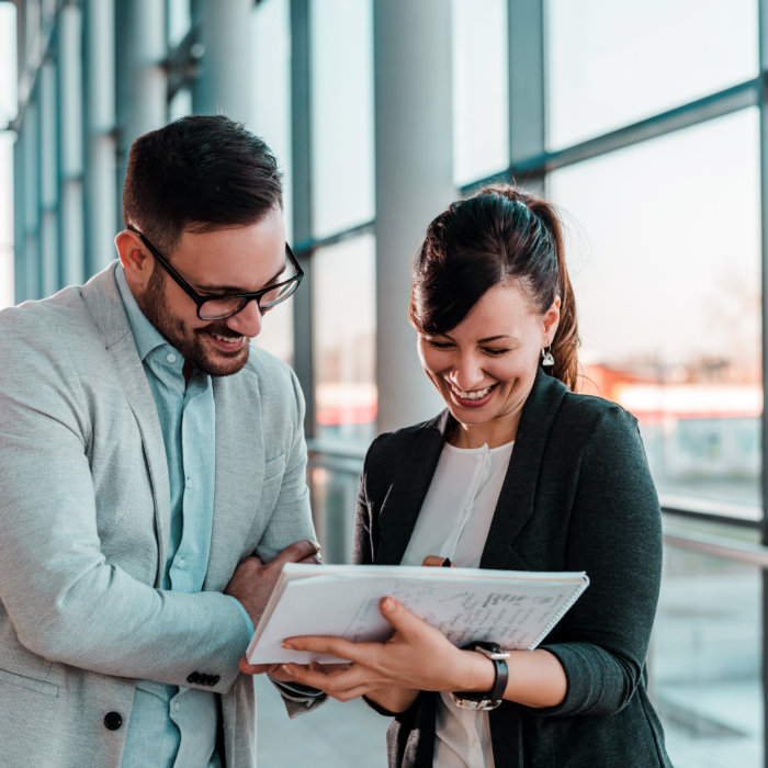 A man and woman are standing together in a bright, modern hallway with floor-to-ceiling windows. Both are smiling and engaged in conversation as they look at a notepad the woman is holding. The man is wearing glasses, a light grey blazer, and a pale blue shirt, with his arms casually crossed. The woman is dressed in a dark blazer over a white blouse and is pointing at something on the notepad. The atmosphere is collaborative and positive, with natural daylight streaming through the windows and reflecting a professional yet relaxed work environment.