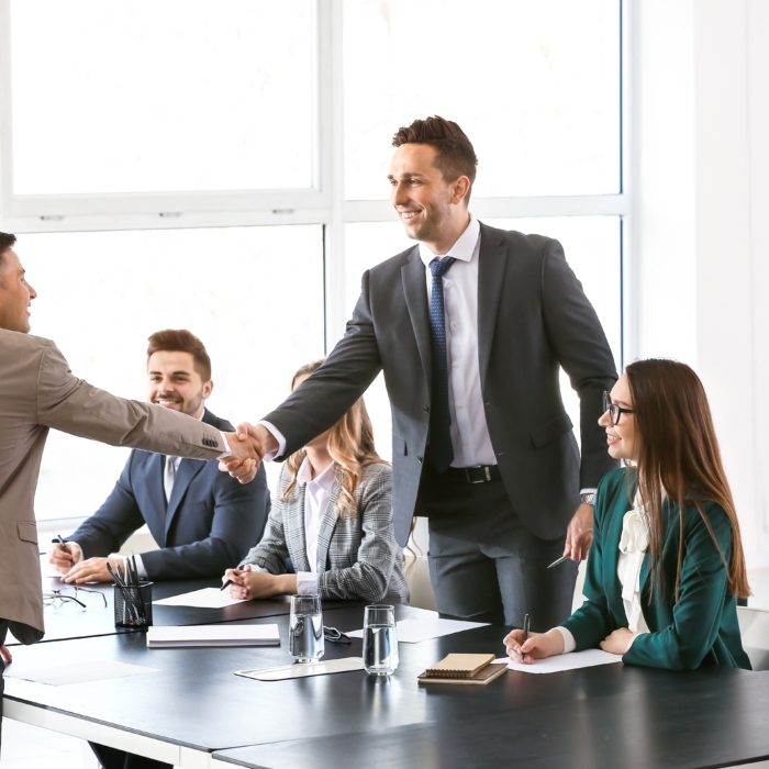 A man in a beige suit shakes hands with a standing interviewer in a dark suit, signaling the beginning or conclusion of a professional meeting or job interview. Both are smiling, indicating a positive and welcoming atmosphere. Around the table, three other individuals—two women and one man—are seated, observing the interaction with interest. Each has a notepad or folder in front of them, suggesting they are part of a panel or team. The setting is a modern, well-lit office with large windows allowing in natural light, contributing to the open and professional tone of the environment.
