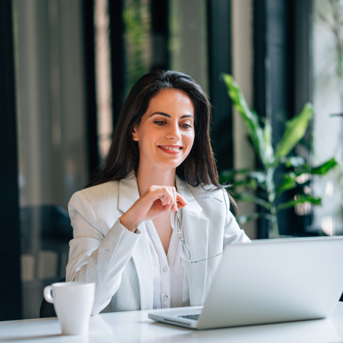 A woman with long dark hair sits at a desk, smiling as she looks at her laptop screen. She is dressed in a light blazer and white blouse, holding a pair of glasses in one hand. A white coffee cup is placed on the desk in front of her. The workspace is modern and well-lit, with large windows and green plants in the background, creating a fresh and professional environment.