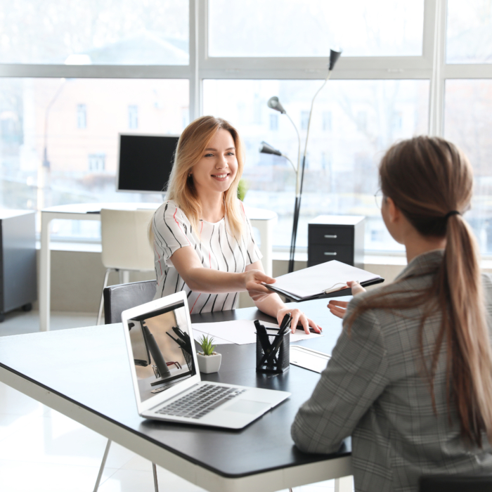 A woman with blonde hair, dressed in a white blouse with vertical stripes, smiles as she hands a document—likely a CV or portfolio—across the table to another woman in a grey plaid blazer. The setting is a bright, modern office, with large windows letting in natural light and offering a view of buildings outside. A man sits beside the woman receiving the document, suggesting a panel or group interview. An open laptop, a small potted plant, and neatly arranged stationery are on the table, creating a clean and professional environment. The interaction appears friendly and engaged, reflecting a positive and collaborative atmosphere.