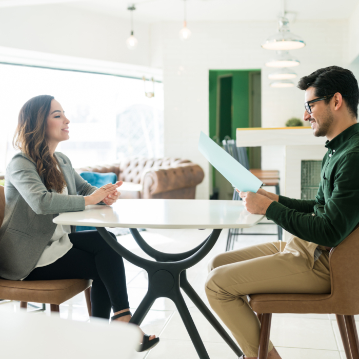 A professional meeting takes place in a bright, modern lounge setting. A woman dressed in a light grey blazer and white blouse sits across from a man in a dark green shirt and beige trousers. The man is holding a folder or document, reviewing its contents with a focused yet approachable expression. The woman maintains engaged body language, seated upright with her hands lightly clasped on the table. The atmosphere suggests a formal yet relaxed discussion, such as a job interview or client consultation. The space features contemporary furnishings, ample natural light, and a clean, professional aesthetic.