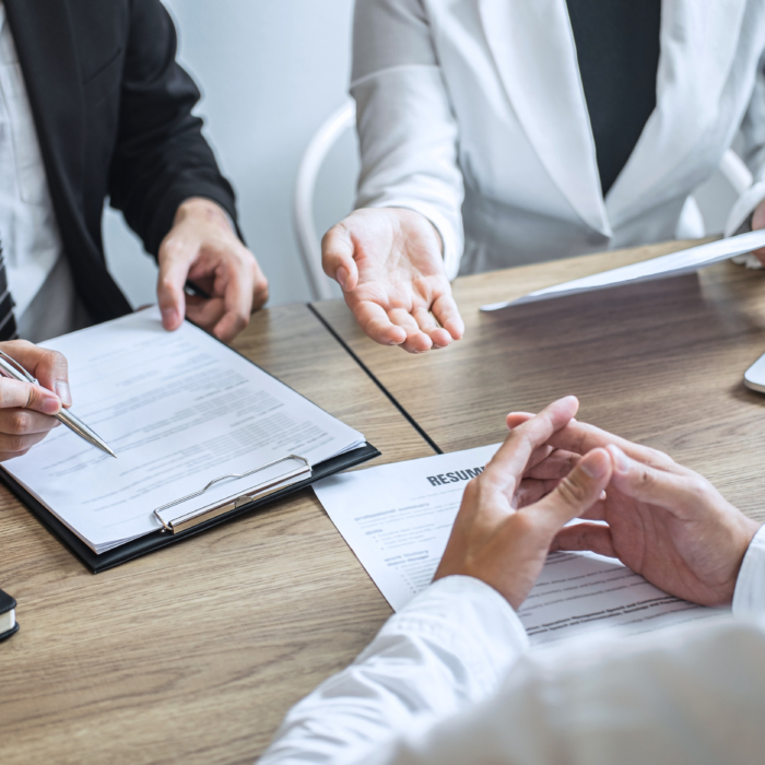 A formal job interview is underway, with two interviewers seated across from a candidate at a conference table. The interviewers are dressed in professional business attire—one in a suit and striped tie, the other in a light blazer—each reviewing documents and making notes. A printed résumé is placed on the table in front of the candidate, who sits with hands folded, listening attentively. A laptop and notepads are visible, indicating a structured, corporate setting focused on assessment and evaluation. The body language suggests a serious yet professional tone, typical of high-stakes recruitment discussions.