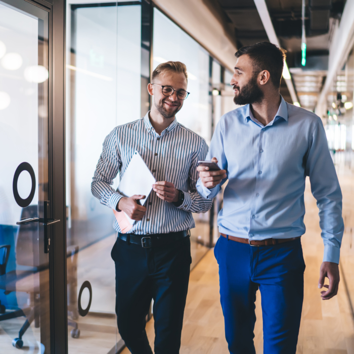 Two professionally dressed men are walking and conversing in a modern office corridor. One is holding a smartphone while the other carries a tablet or folder. Both are smiling and engaged in conversation. The office features glass walls with circular decals, wooden flooring, and a contemporary, open-plan design with visible meeting rooms. The setting conveys a professional, collaborative work environment.