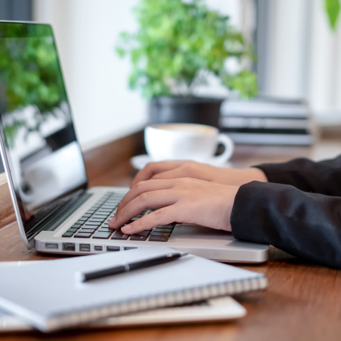 A professional seated at a wooden desk types on a laptop in a clean, well-lit workspace. The scene includes a neatly arranged notebook with a pen, a digital tablet, and a cup of coffee in the background, creating a focused and organised environment. Green potted plants add a touch of natural decor, while the soft daylight enhances the calm and productive atmosphere. The individual is dressed in business attire, suggesting remote work, independent project work, or virtual collaboration in a modern business setting.