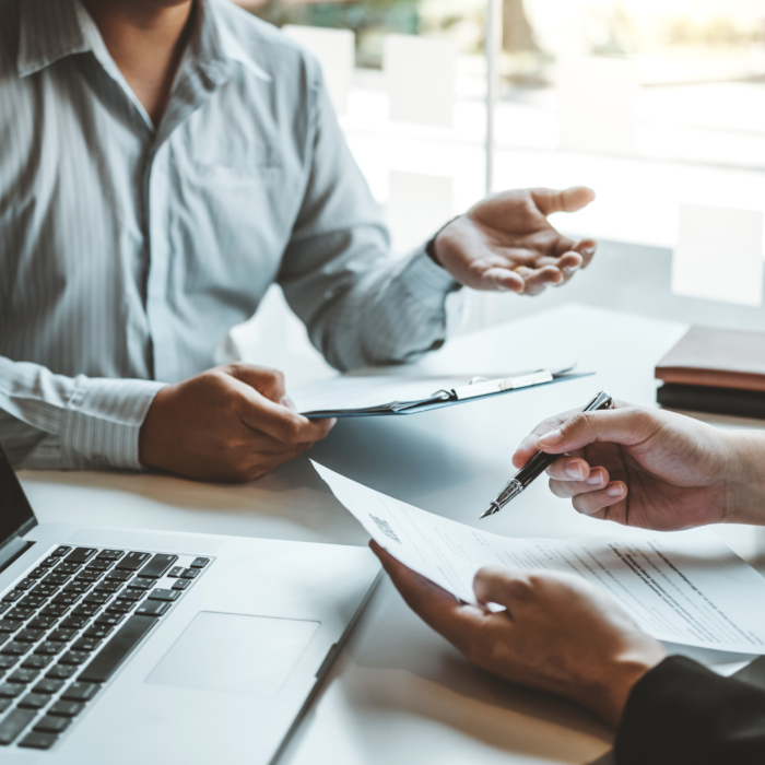 Two people are seated at a table, likely in a business setting. 

One person, wearing a light gray, button-down shirt, is holding a folder or documents. The other person, wearing a dark blazer, is holding a pen and reviewing documents placed on the table. 

A laptop computer is visible on the table between them. The documents appear to be forms or contracts. The setting is an office environment, with a slightly blurred background that suggests a window.