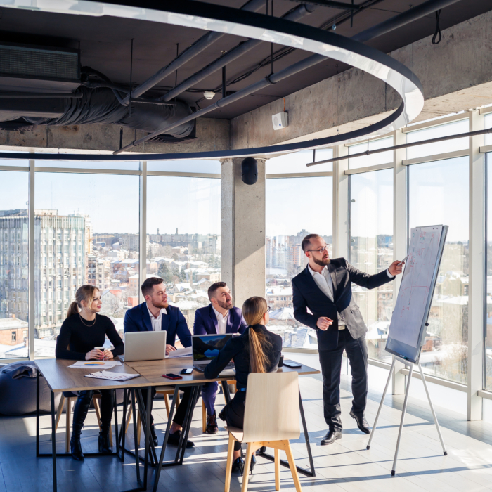 A group of business professionals are gathered in a modern, bright office space for a presentation. 

A man in a dark suit is standing at a whiteboard, pointing to it, leading a presentation to a group of colleagues. The group is seated around a light-colored wooden table, and several individuals are seated at the table with laptops open while others are standing.

The room features large windows overlooking a cityscape, and a large, circular ceiling fixture hangs above. The space is brightly lit and airy, suggesting a modern office environment. The layout is designed for efficient meetings and presentations.