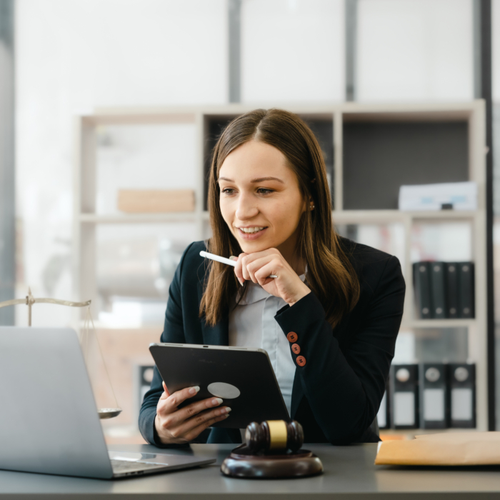 A professional woman sits at a desk in a modern office environment, dressed in business attire. She holds a digital tablet in one hand and a pen in the other, appearing focused and engaged while looking at her laptop screen. A wooden gavel, legal scale, and envelope are placed on the desk, indicating a legal or judicial setting. The background features shelves with files and documents, reinforcing the formal and structured nature of the workspace. The scene conveys a sense of professionalism, concentration, and informed decision-making.