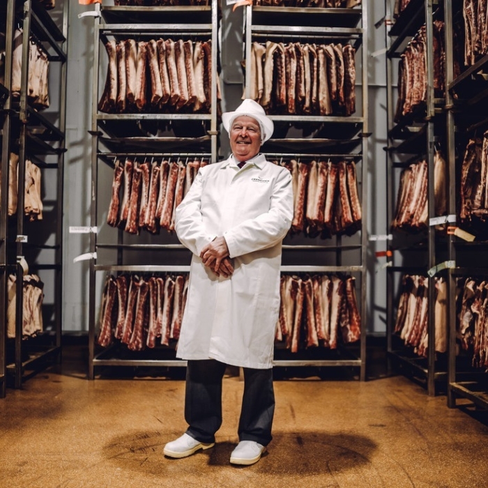 A Cranswick employee proudly standing in front of hanging racks of dry-cured bacon, representing craftsmanship, quality, and experience in food production.