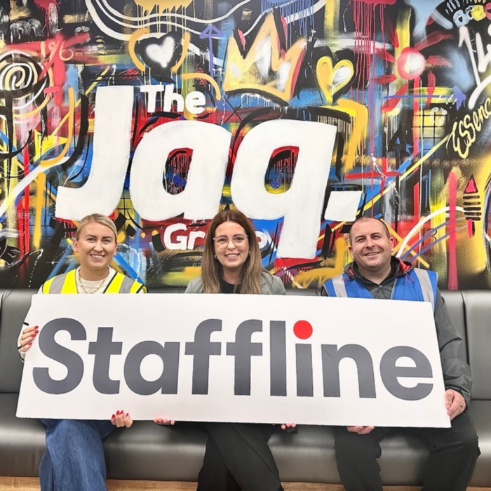 Three people are seated on a grey bench in front of a colourful graffiti-style mural featuring the words “The Jaq Group.” They are smiling and holding a large Staffline sign with the company’s logo in grey and red. The person on the left is wearing a yellow high-visibility vest, the person in the middle is dressed in business attire, and the person on the right is wearing a blue high-visibility vest. The vibrant mural behind them includes abstract patterns, bright colours, and the word “Essence.” The overall image conveys a friendly and collaborative partnership between Staffline Recruitment Ireland and The Jaq Group.