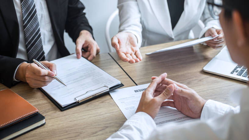 A formal job interview is underway, with two interviewers seated across from a candidate at a conference table. The interviewers are dressed in professional business attire—one in a suit and striped tie, the other in a light blazer—each reviewing documents and making notes. A printed résumé is placed on the table in front of the candidate, who sits with hands folded, listening attentively. A laptop and notepads are visible, indicating a structured, corporate setting focused on assessment and evaluation. The body language suggests a serious yet professional tone, typical of high-stakes recruitment discussions.