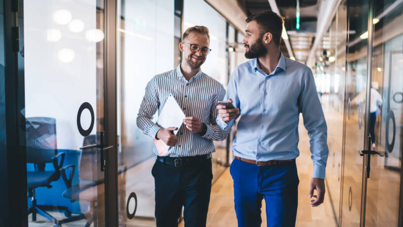 Two professionally dressed men are walking and conversing in a modern office corridor. One is holding a smartphone while the other carries a tablet or folder. Both are smiling and engaged in conversation. The office features glass walls with circular decals, wooden flooring, and a contemporary, open-plan design with visible meeting rooms. The setting conveys a professional, collaborative work environment.