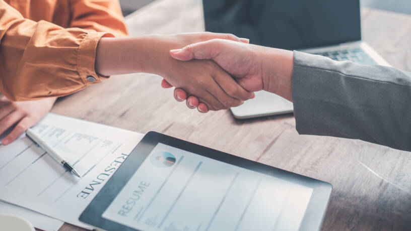 Two people shake hands across a desk, signalling the successful conclusion of a job interview or meeting. On the table are a printed CV, a tablet displaying a digital version of a resume, and an open laptop, reflecting a modern, hybrid approach to recruitment. One person is wearing an orange shirt, while the other is in a grey blazer, suggesting a formal but approachable setting. The close-up focus on the handshake captures a moment of agreement or a potential job offer, emphasising professionalism, trust, and mutual respect.