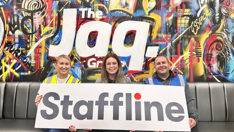 Three people are seated on a grey bench in front of a colourful graffiti-style mural featuring the words “The Jaq Group.” They are smiling and holding a large Staffline sign with the company’s logo in grey and red. The person on the left is wearing a yellow high-visibility vest, the person in the middle is dressed in business attire, and the person on the right is wearing a blue high-visibility vest. The vibrant mural behind them includes abstract patterns, bright colours, and the word “Essence.” The overall image conveys a friendly and collaborative partnership between Staffline Recruitment Ireland and The Jaq Group.